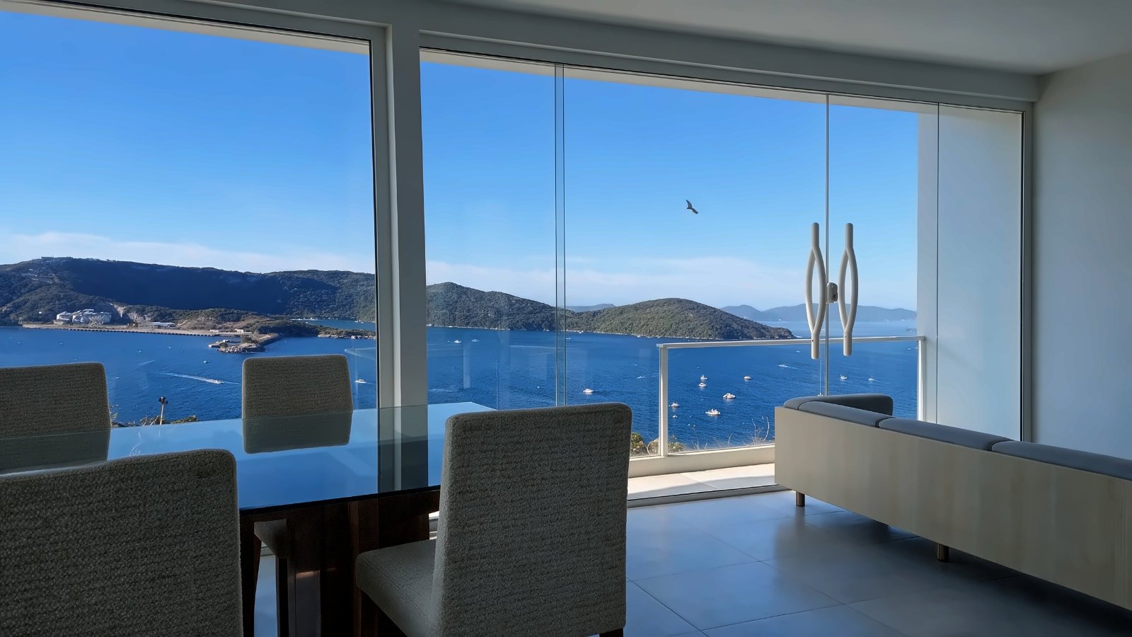 Sala com janelas do chão ao teto voltadas para a enseada do Pontal do Atalaia, com barcos e a ilha ao fundo